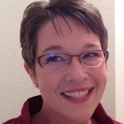 Close-up portrait of a smiling woman with short hair and glasses against a light-colored wall.