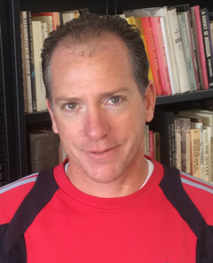Head-and-shoulders portrait of a man in a red and black sweatshirt, standing in front of a bookshelf filled with books.