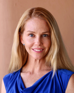 Smiling woman with long blonde hair wearing a blue sleeveless top, posed against a warm tan background.