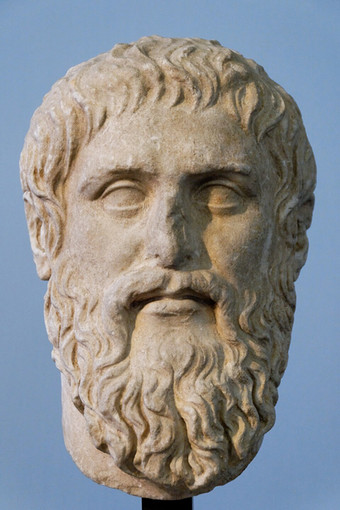 Close-up of an ancient marble sculpture head of a bearded man with curly hair, set against a blue background.