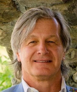 Head-and-shoulders portrait of a smiling man with swept-back gray hair, photographed outdoors beside a stone wall.